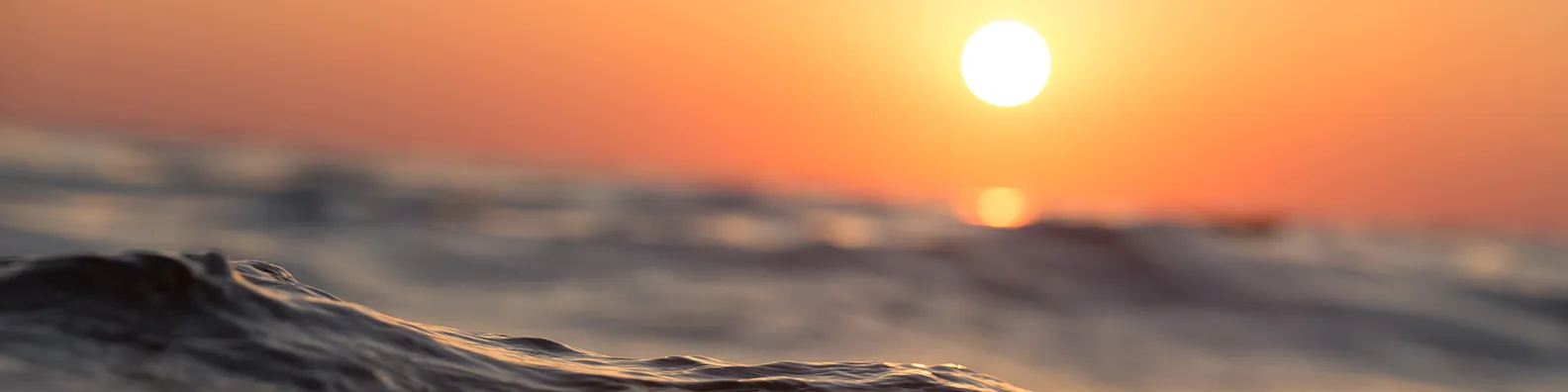 Sunset over the ocean with waves and a bright sun in the sky.