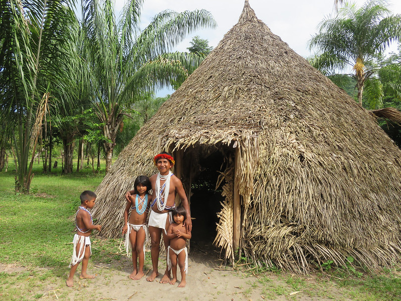 Indigenous community members standing outside traditional thatched hut in lush environment.