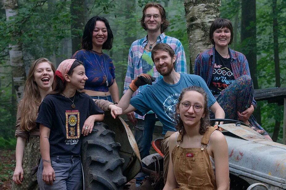 Group of diverse young adults smiling outdoors with tractor in a forest setting.