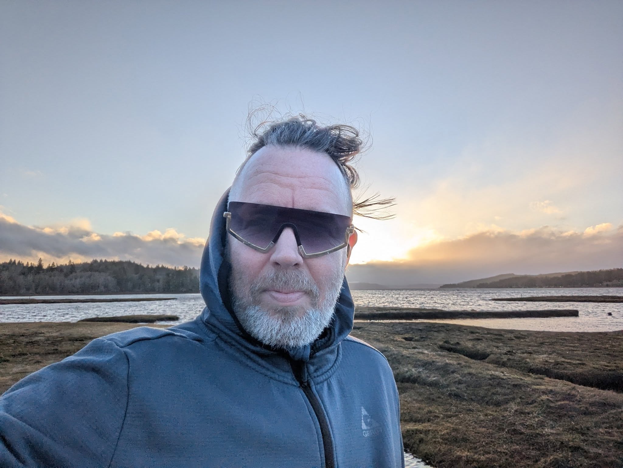 Man in sunglasses outdoors at sunset near a river with mountains in the background.