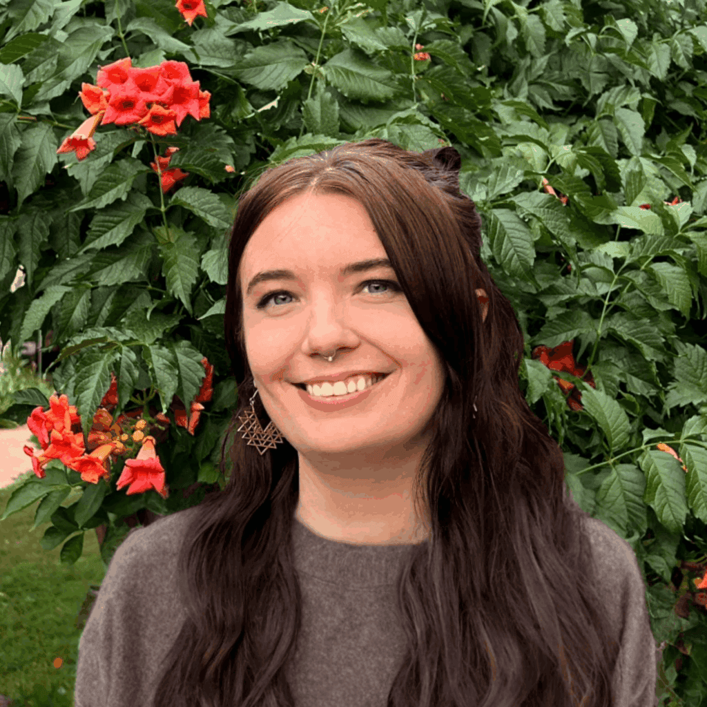 Young woman smiling in a lush garden with red flowers and green foliage.