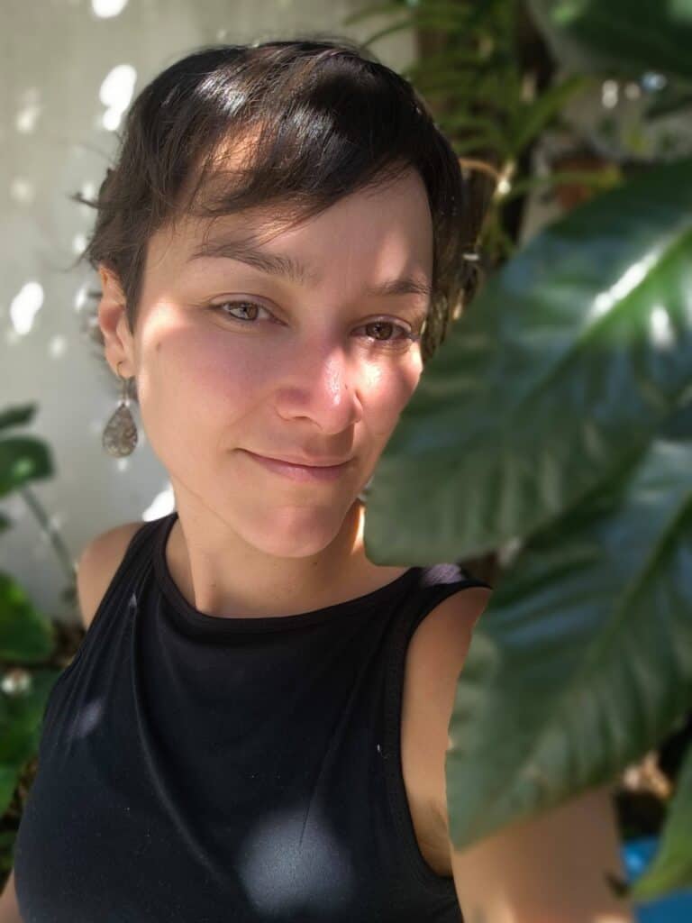 Portrait of a woman with short dark hair and earrings in a lush green setting.