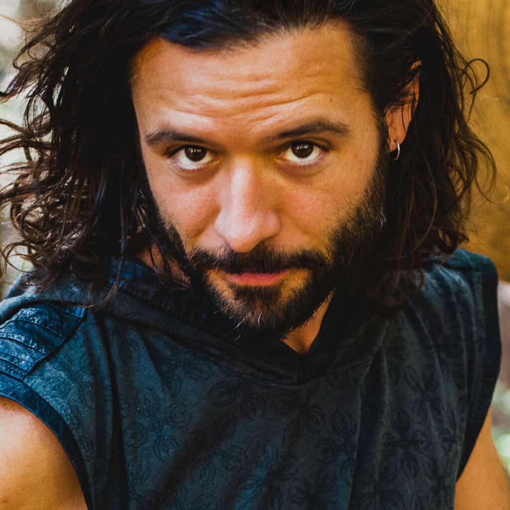 Close-up portrait of a man with long curly hair and beard, looking directly at the camera.