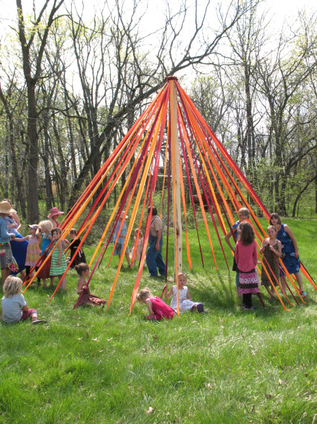 Children playing around a colorful outdoor maypole in a green park.